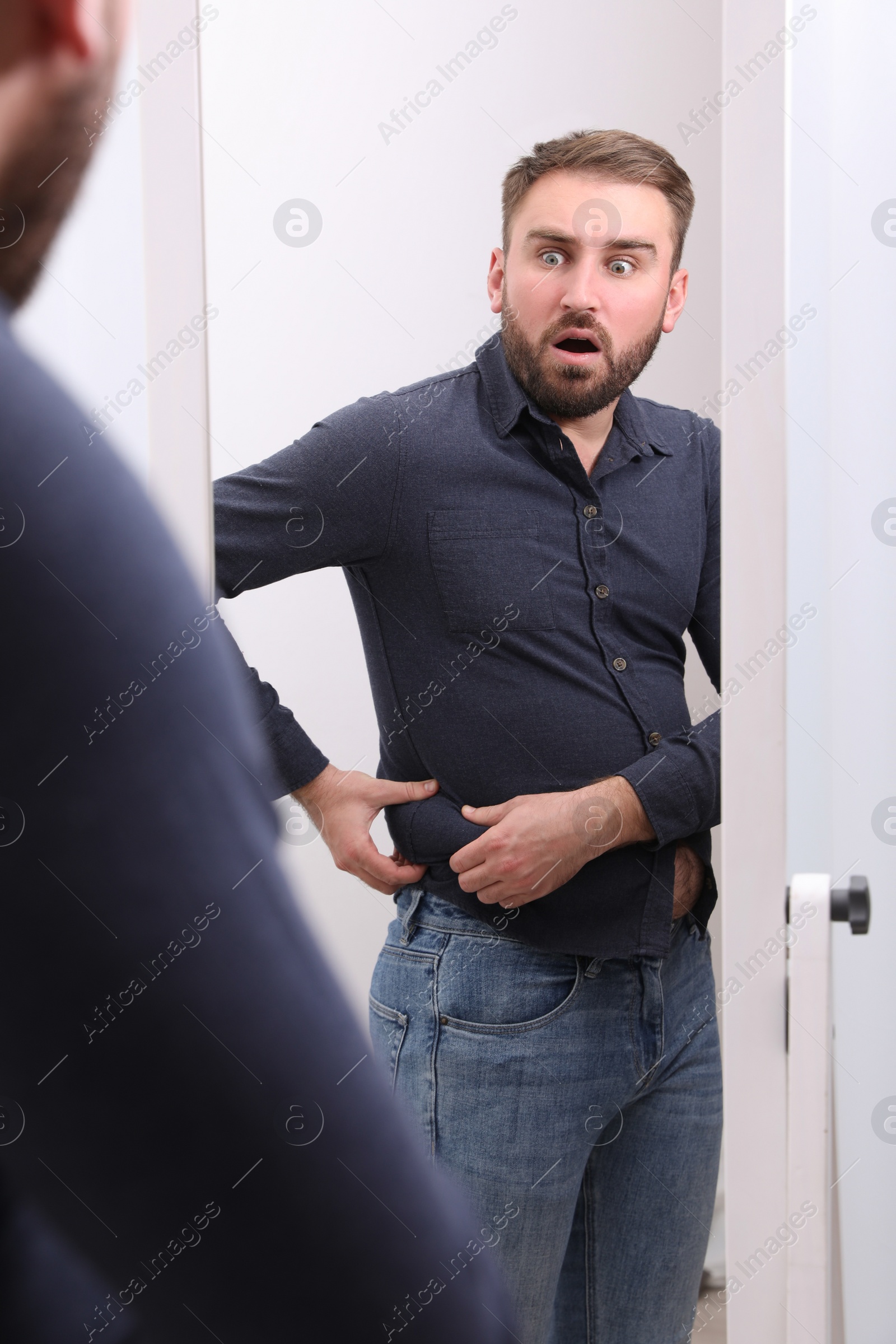 Photo of Scared man wearing tight shirt in front of mirror at home. Overweight problem