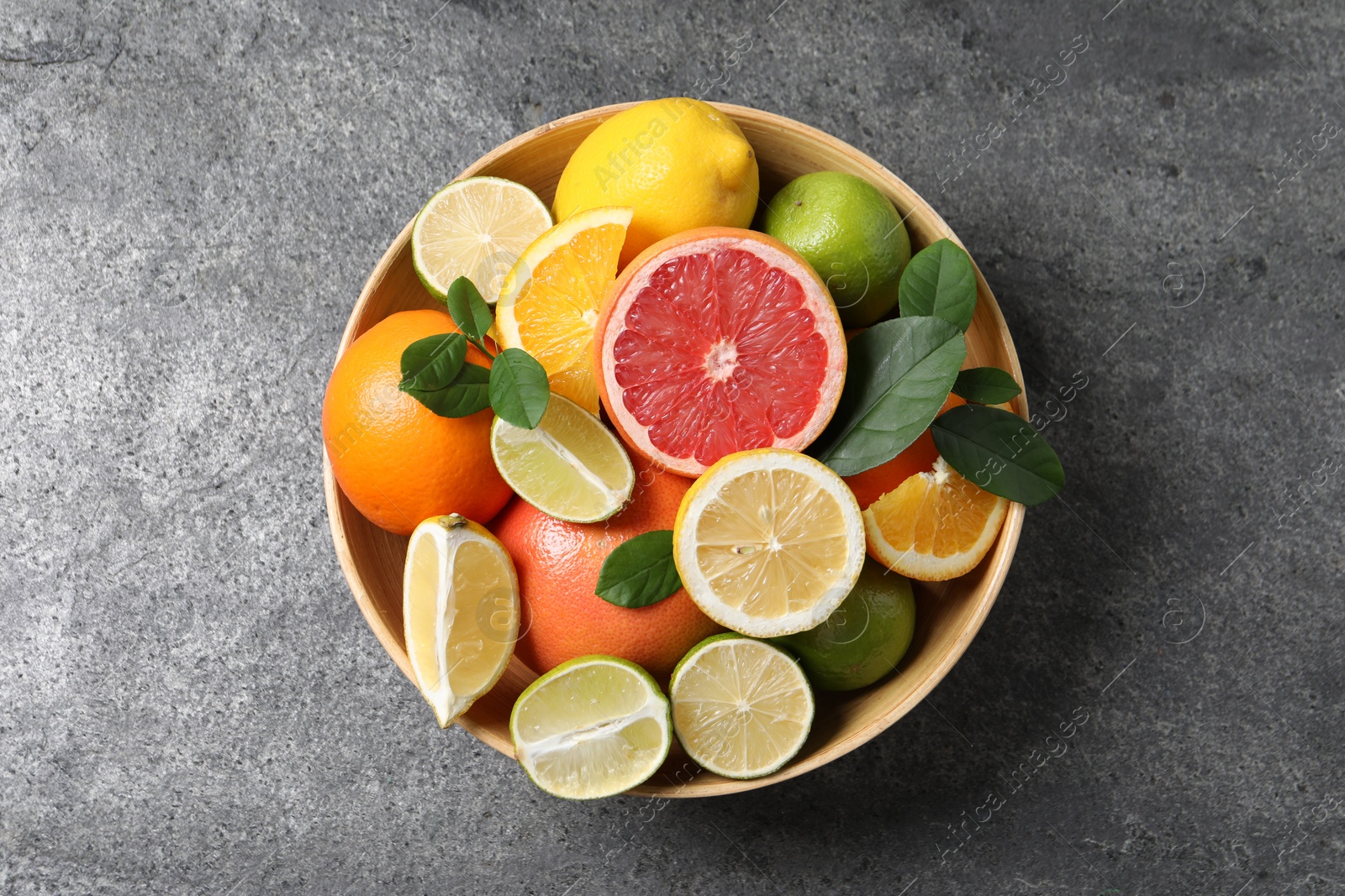 Photo of Different fresh citrus fruits and leaves in bowl on grey textured table, top view