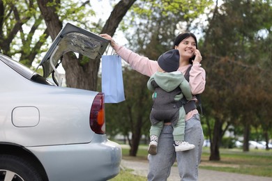 Mother holding her child in sling (baby carrier) while closing car trunk outdoors