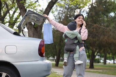 Photo of Mother holding her child in sling (baby carrier) while closing car trunk outdoors