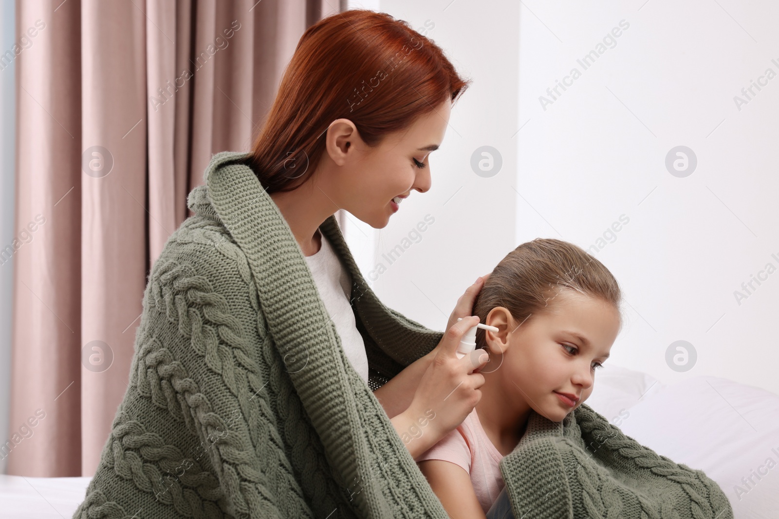 Photo of Mother spraying medication into daughter's ear while being wrapped in warm plaid at home