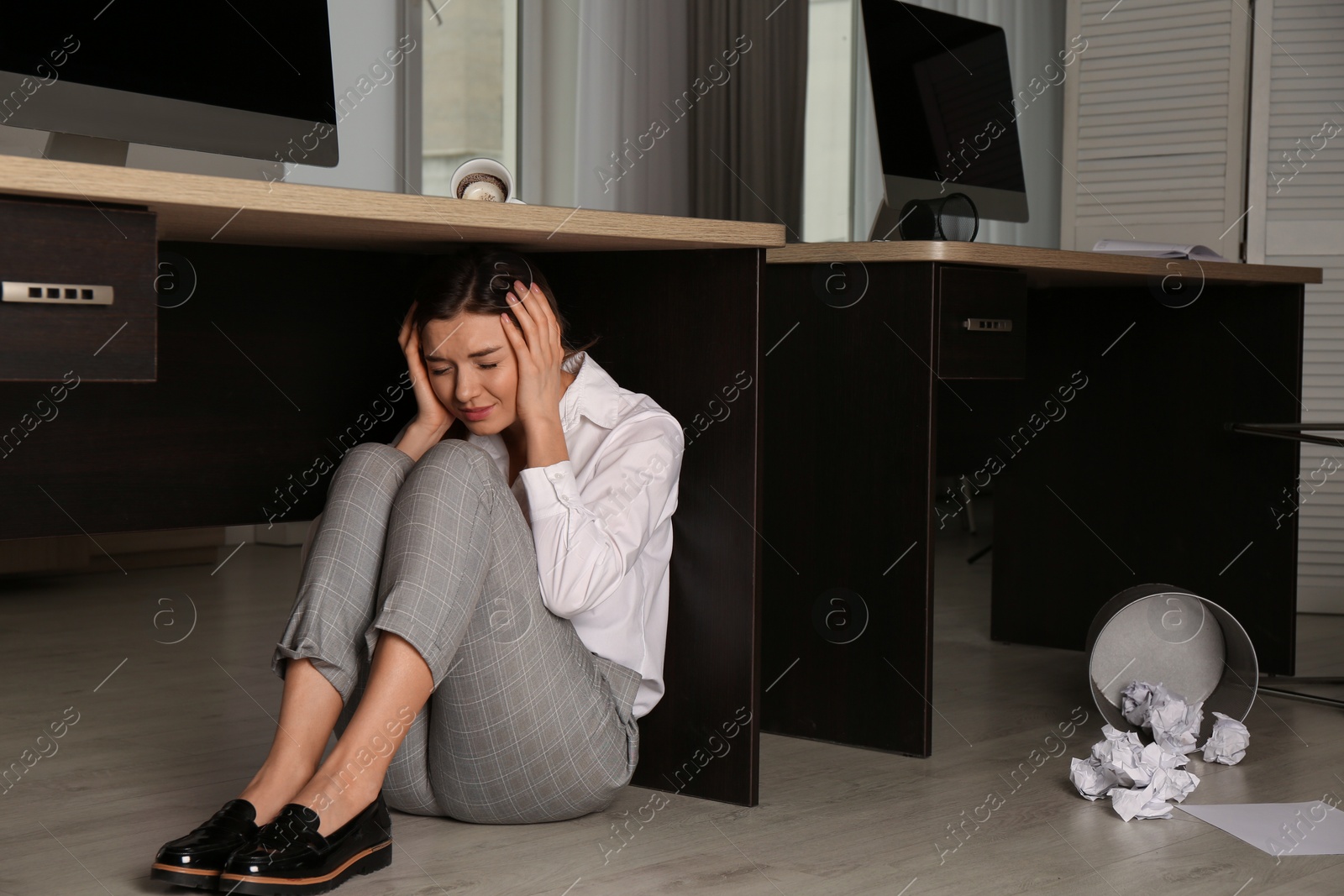 Photo of Scared young woman hiding under office desk during earthquake