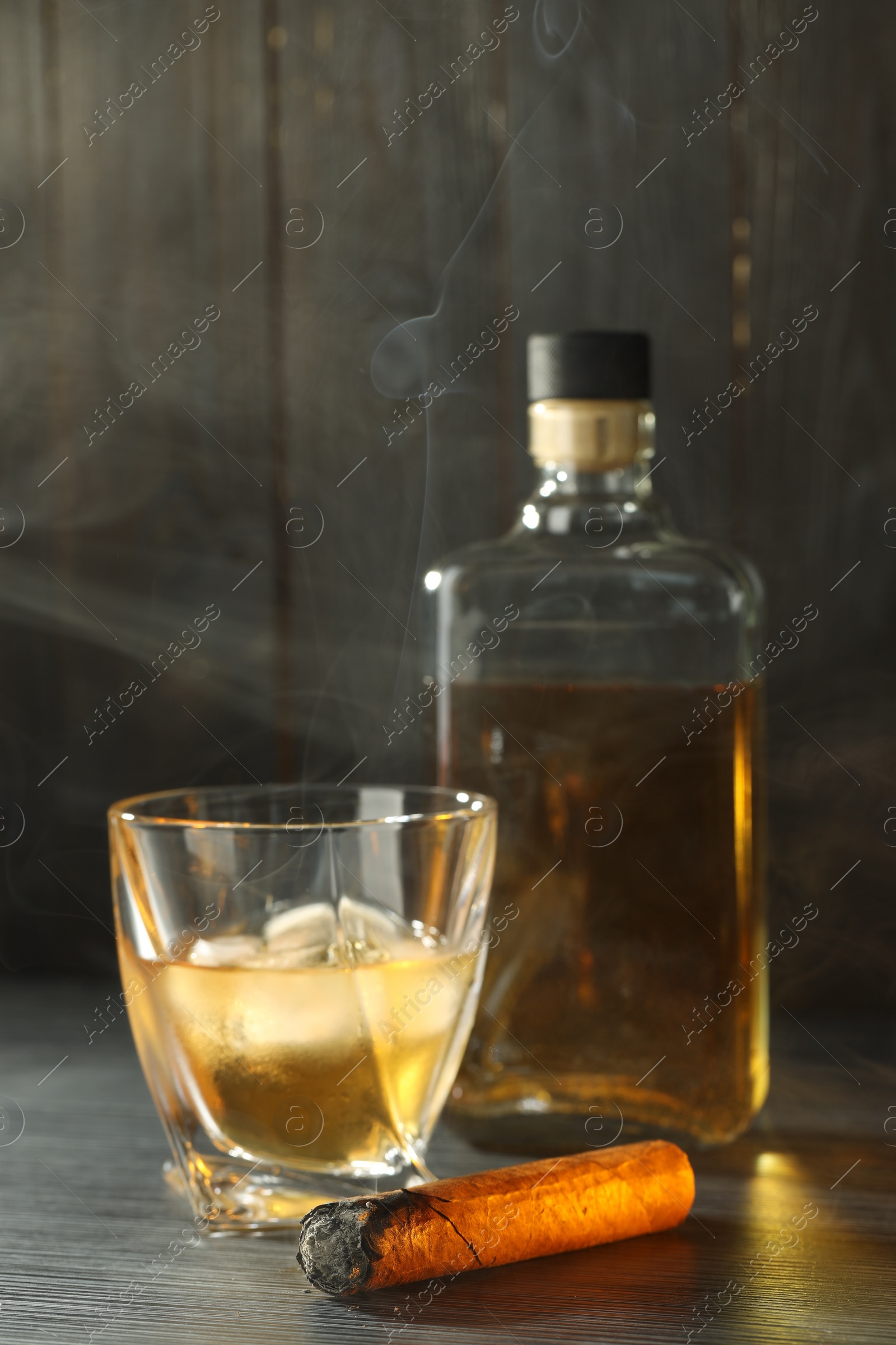 Photo of Bottle, glass of whiskey with ice cubes and smoldering cigar on black wooden table