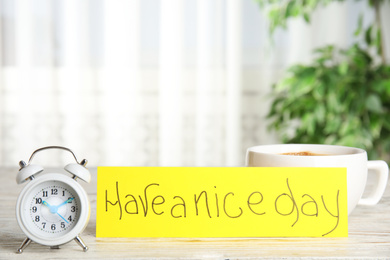 Photo of Delicious morning coffee, alarm clock and card with HAVE A NICE DAY wish on white wooden table indoors