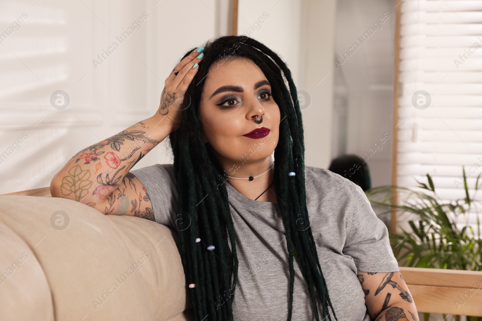 Photo of Beautiful young woman with tattoos on body, nose piercing and dreadlocks at home