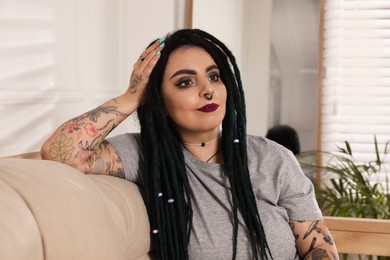 Photo of Beautiful young woman with tattoos on body, nose piercing and dreadlocks at home