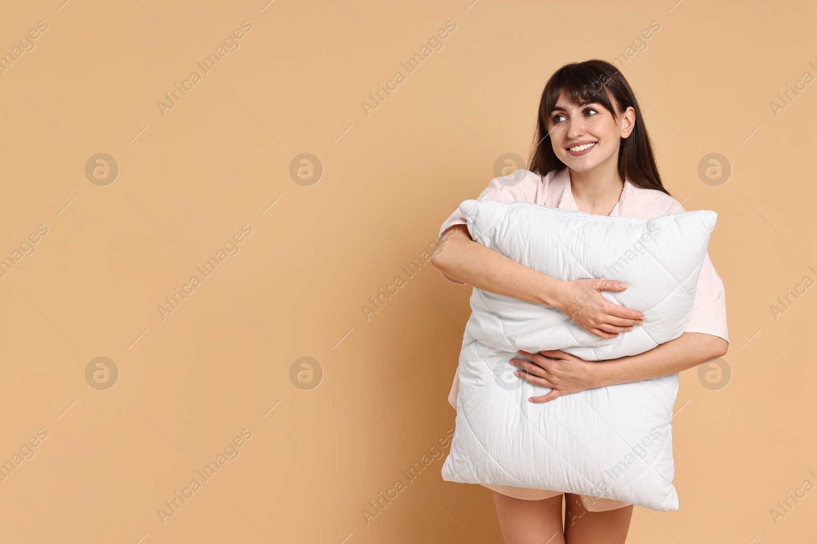 Photo of Happy woman wearing pyjama and holding pillow on beige background, space for text