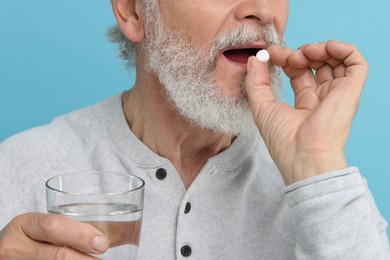 Senior man with glass of water taking pill on light blue background, closeup