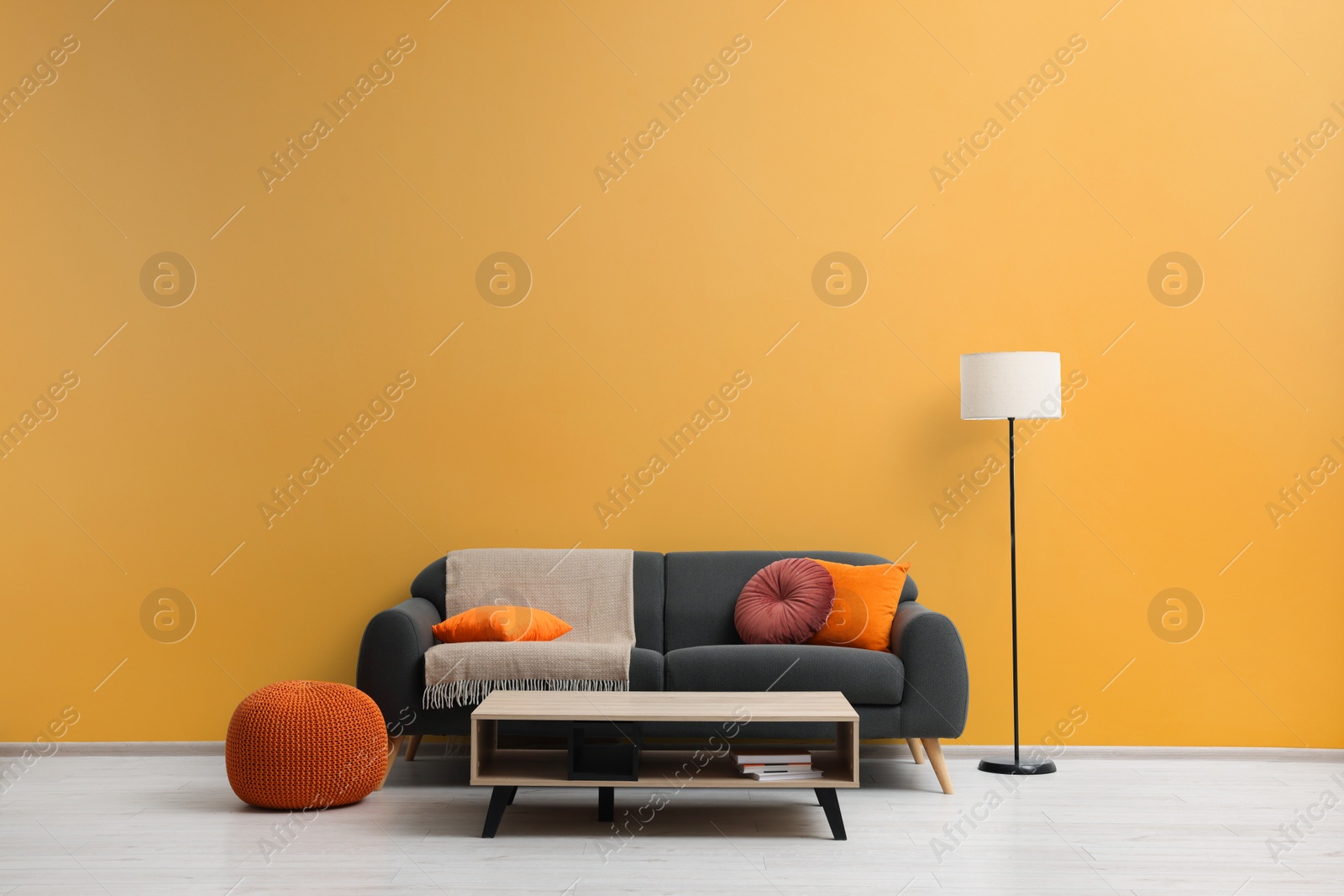 Photo of Stylish grey sofa with colorful pillows, wooden table and lamp near pale orange wall indoors. Interior design