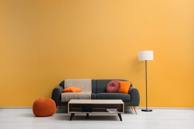Photo of Stylish grey sofa with colorful pillows, wooden table and lamp near pale orange wall indoors. Interior design