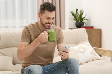 Photo of Happy man holding delicious fresh smoothie and using smartphone at home