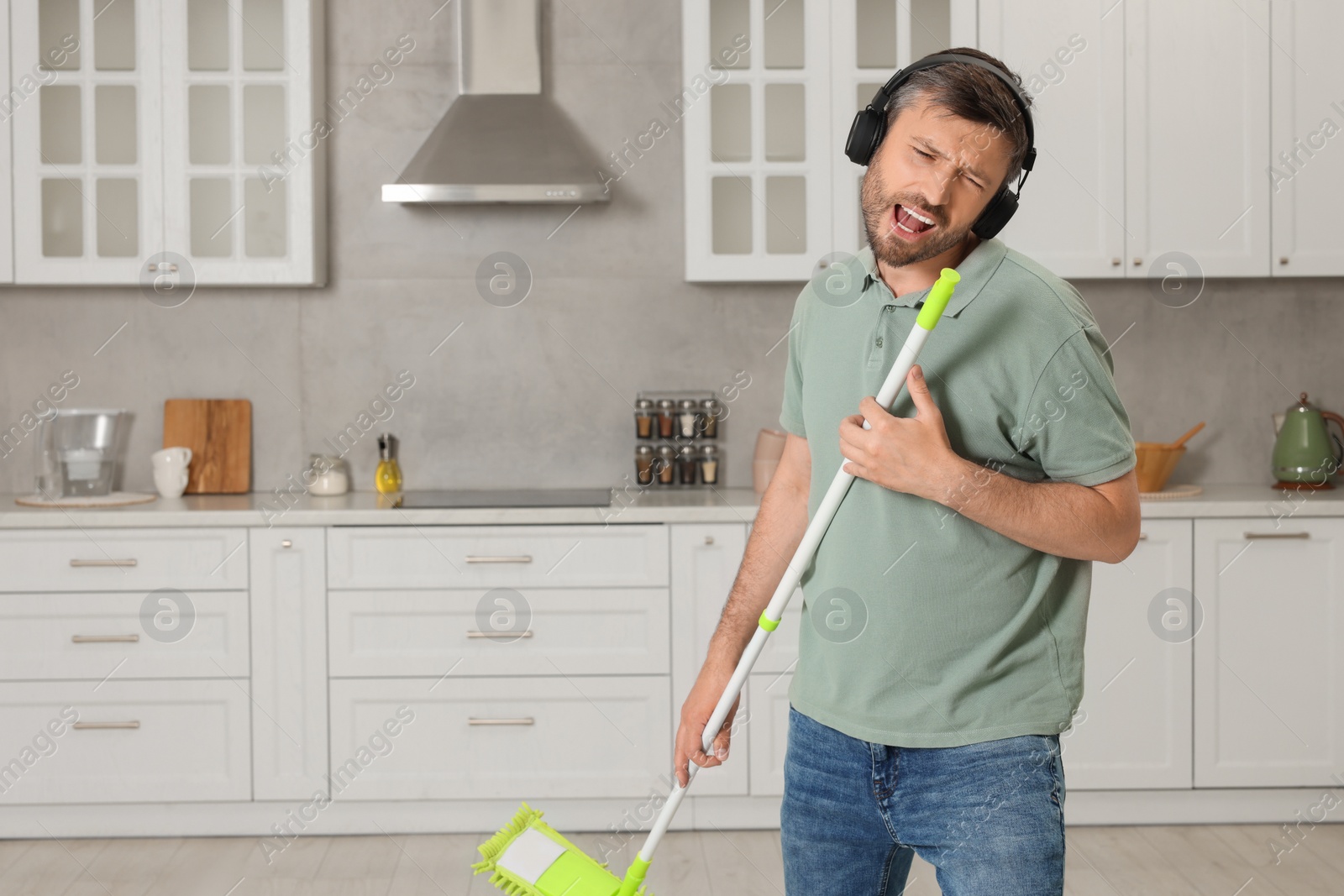 Photo of Happy man in headphones with mop singing while cleaning in kitchen. Space for text