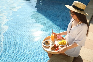 Photo of Young woman with delicious breakfast on tray near swimming pool. Space for text