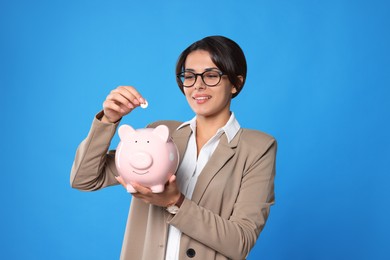 Young woman putting coin into piggy bank on light blue background