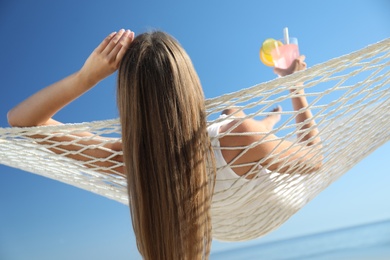 Photo of Young woman with refreshing cocktail relaxing in hammock on beach