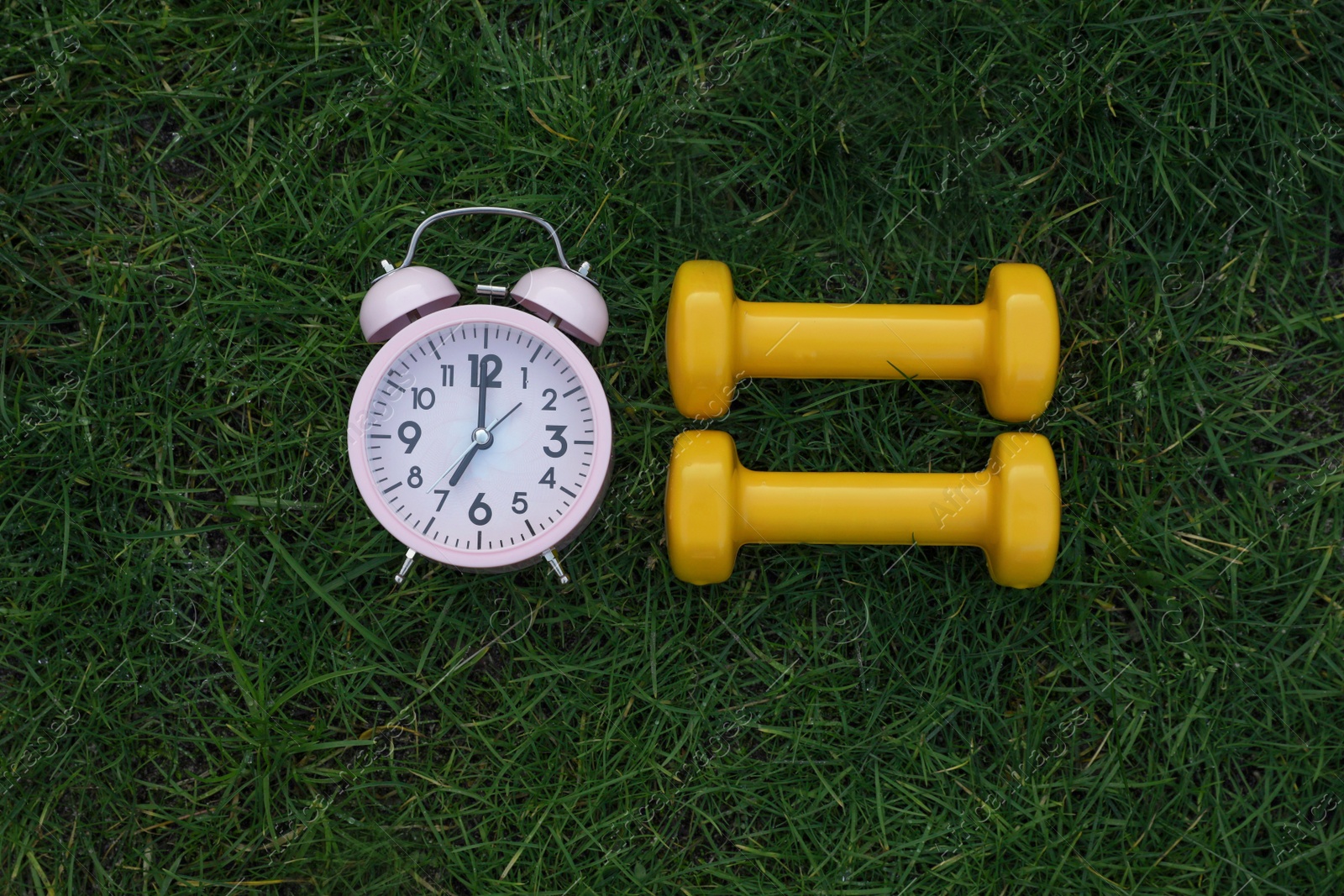 Photo of Alarm clock and dumbbells on green grass, flat lay. Morning exercise