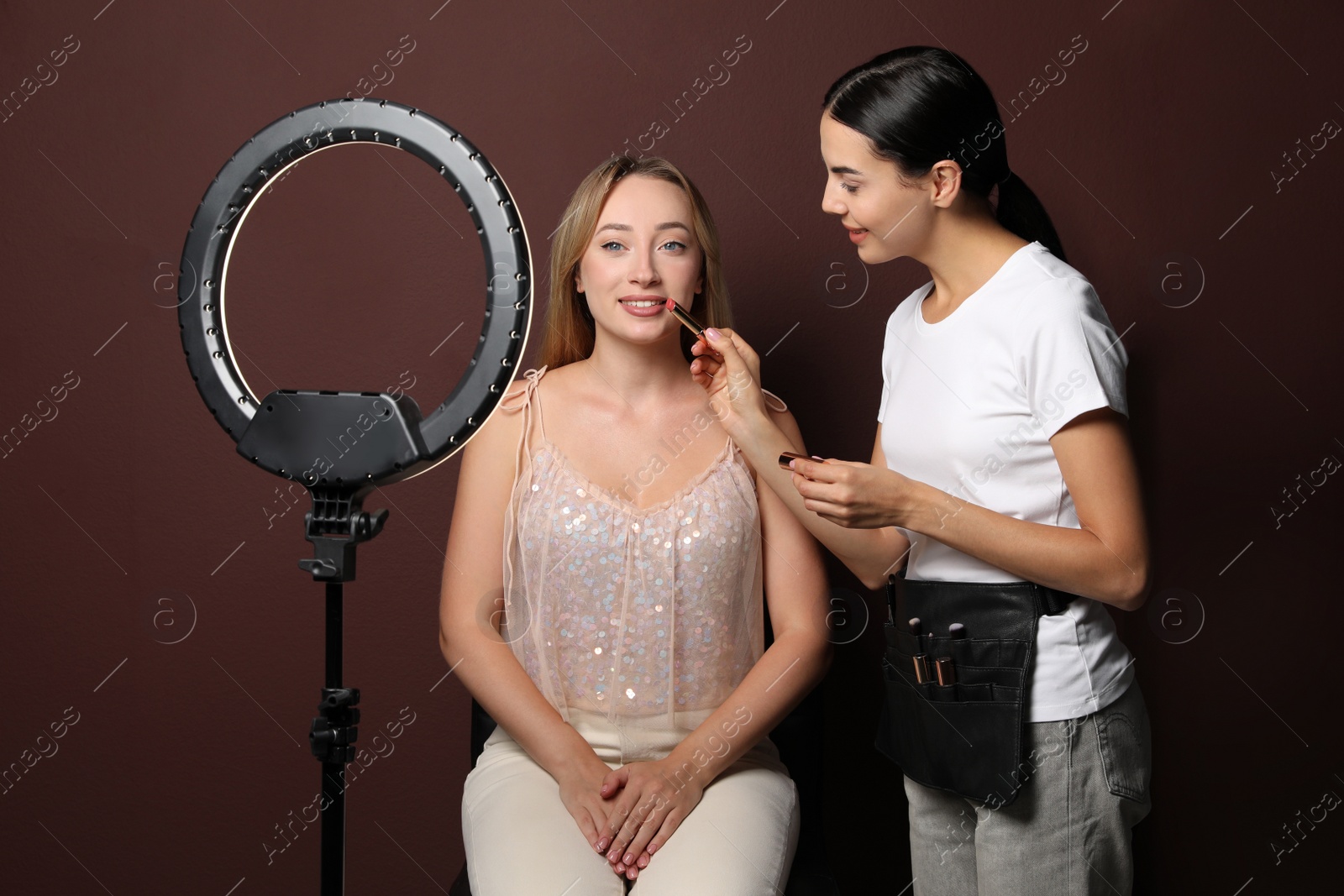 Photo of Professional makeup artist working with beautiful young woman against brown background. Using ring lamp