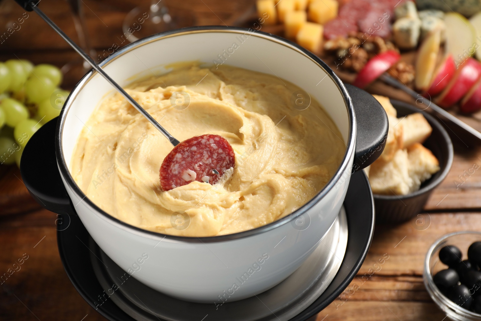 Photo of Fork with piece of sausage and melted cheese in fondue pot on wooden table, closeup