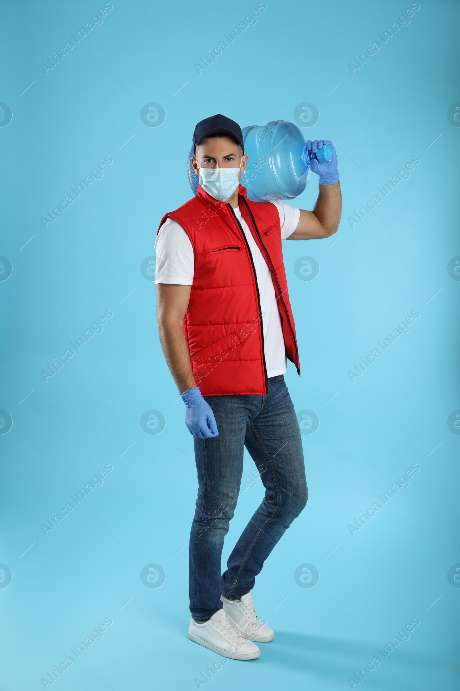 Photo of Courier in face mask with bottle of cooler water on light blue background. Delivery during coronavirus quarantine