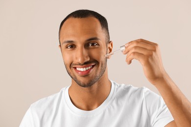 Handsome man applying cosmetic serum onto face on light grey background