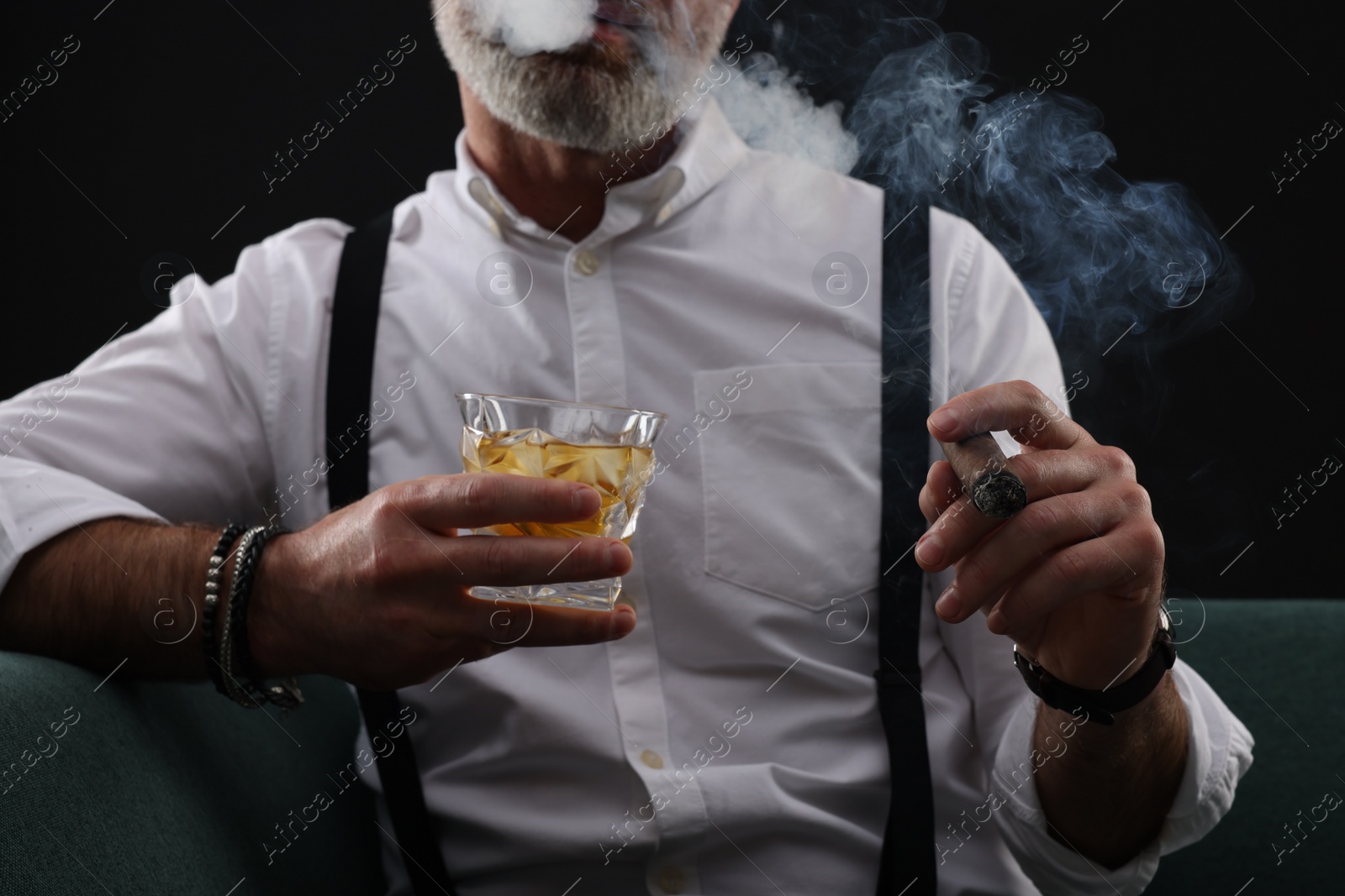 Photo of Man with glass of whiskey smoking cigar on black background, closeup