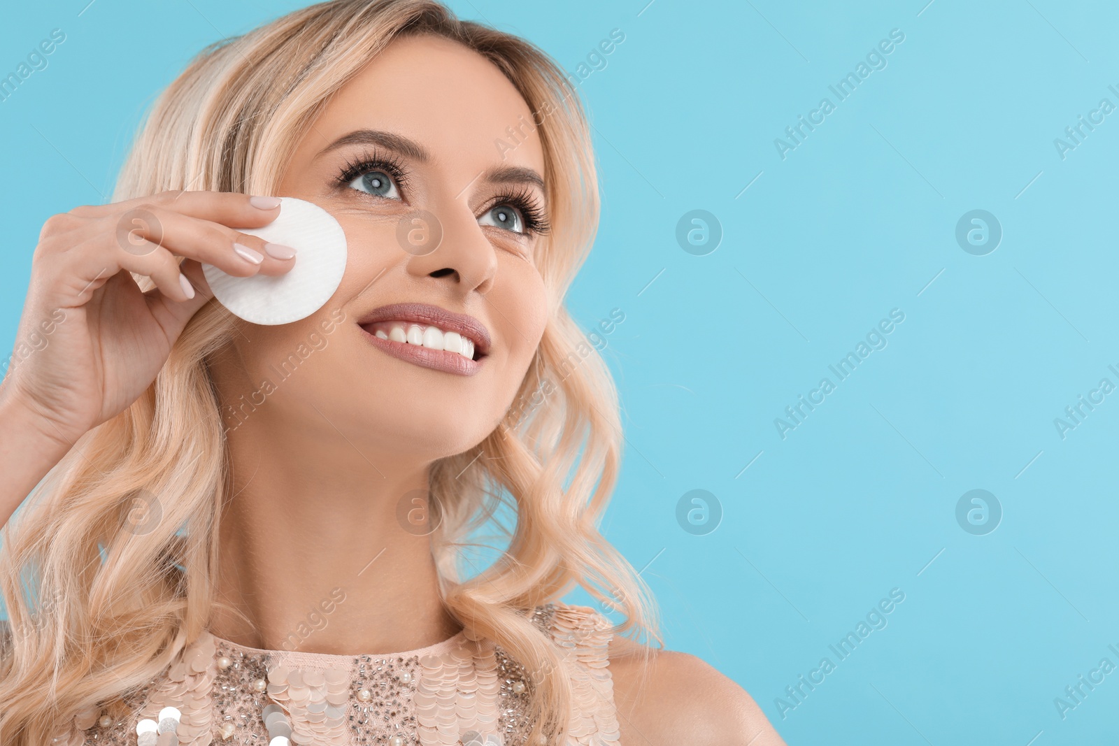 Photo of Smiling woman removing makeup with cotton pad on light blue background, closeup. Space for text