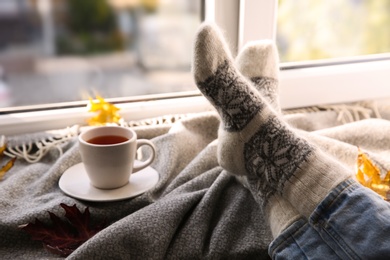 Woman in knitted socks relaxing with cup of hot drink and plaid near window at home, closeup