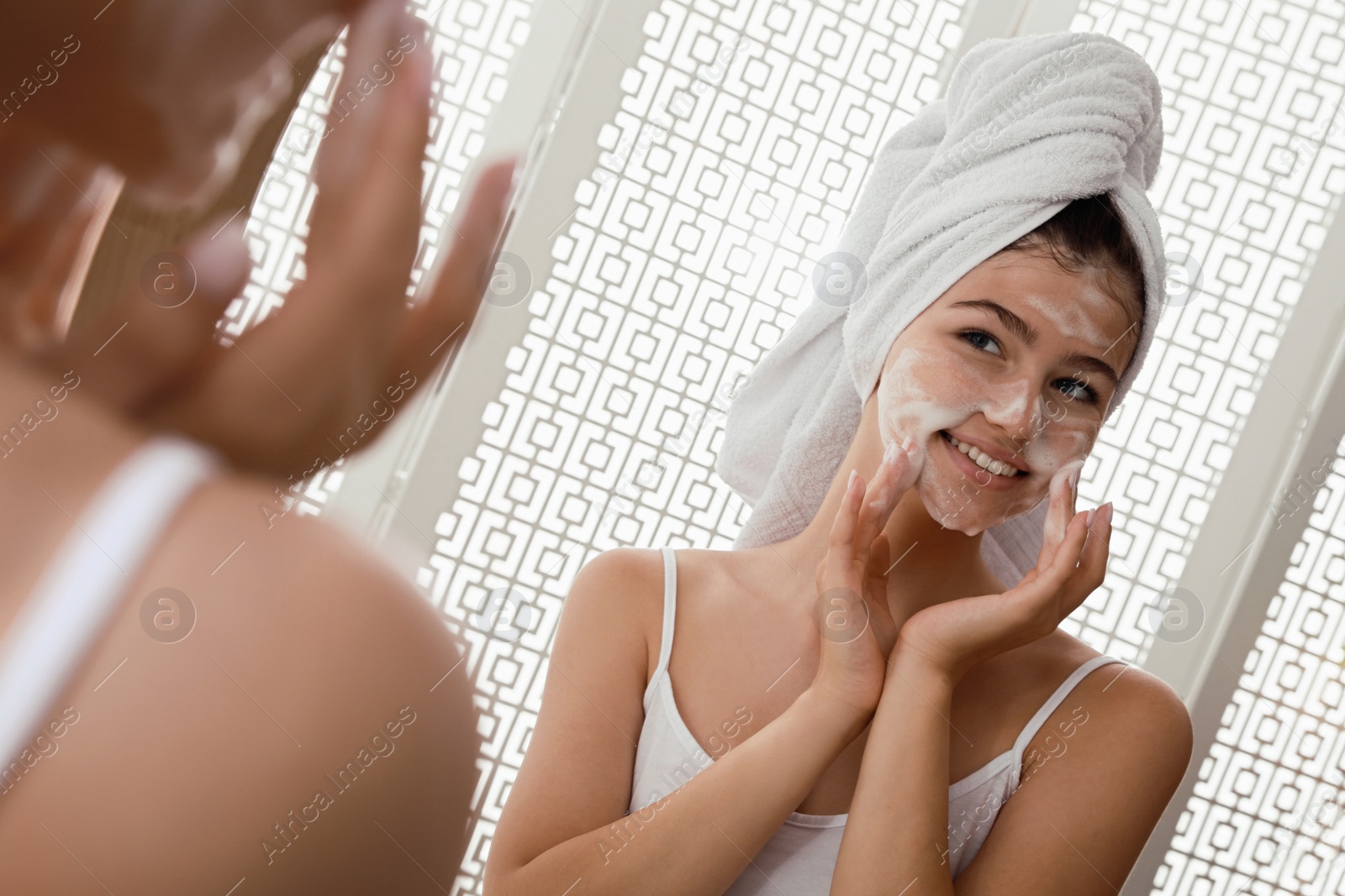 Photo of Beautiful teenage girl applying cleansing foam onto face near mirror at home. Skin care cosmetic