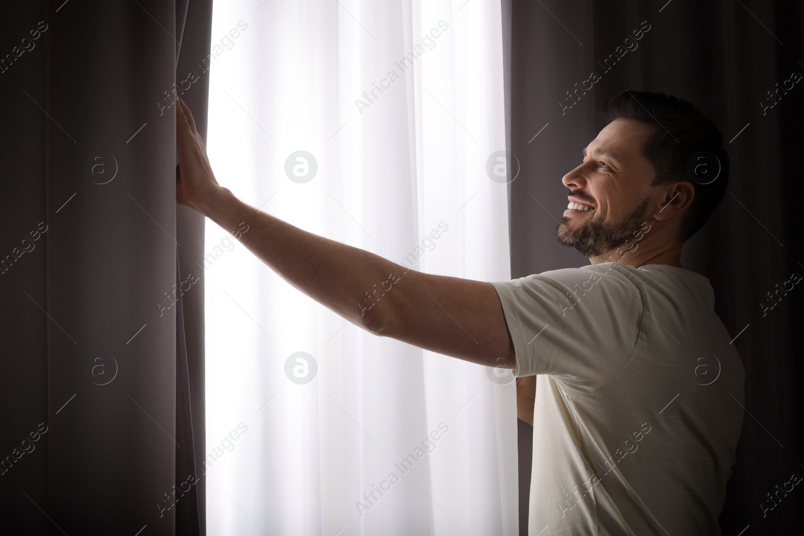 Photo of Happy man opening window curtains at home