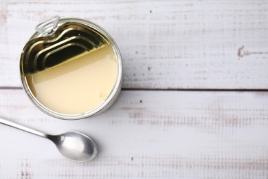Photo of Tasty condensed milk in tin can and spoon on white wooden table, top view. Space for text