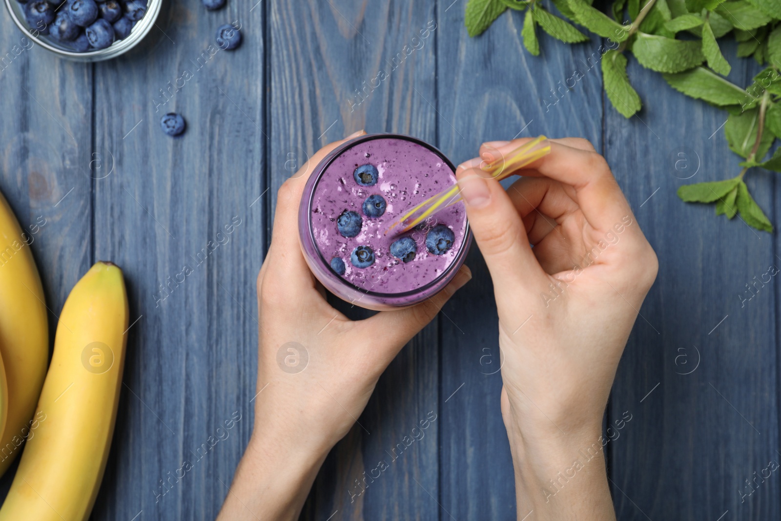 Photo of Woman holding glass of delicious blueberry smoothie at blue wooden table, top view