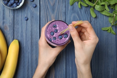 Woman holding glass of delicious blueberry smoothie at blue wooden table, top view