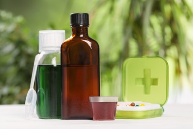 Photo of Bottles of syrup, measuring cup, dosing spoon and pills on white table against blurred background. Cold medicine
