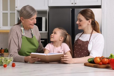 Photo of Cute little girl with her mother and grandmother cooking by recipe book in kitchen