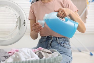 Photo of Woman pouring fabric softener from bottle into cap near washing machine in bathroom, closeup