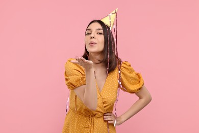 Photo of Young woman in party hat blowing kiss on pink background