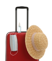 Photo of Stylish red suitcase with hat, antiseptic spray and protective mask on white background. Travelling during coronavirus pandemic