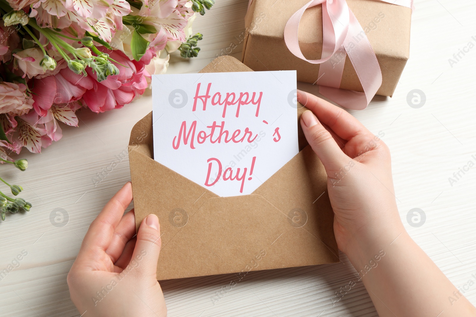 Image of Happy Mother's Day. Woman taking greeting card out of envelope, closeup. Gift box and bouquet of beautiful flowers on white wooden table