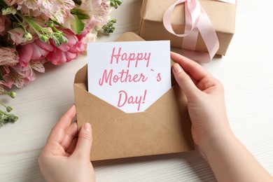 Happy Mother's Day. Woman taking greeting card out of envelope, closeup. Gift box and bouquet of beautiful flowers on white wooden table