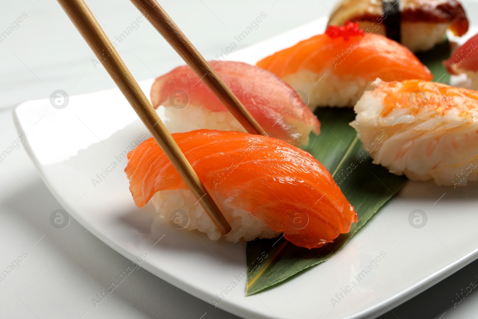 Photo of Taking delicious nigiri sushi with chopsticks from plate on white table, closeup