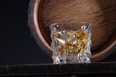 Whiskey with ice cubes in glass and barrel on wooden table against black background, closeup. Space for text