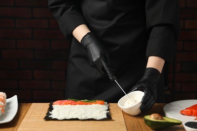 Photo of Chef in gloves putting cream cheese onto unwrapped sushi roll at wooden table, closeup