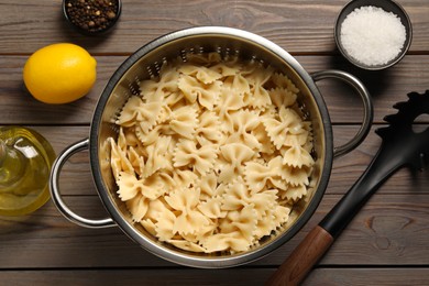 Cooked pasta in metal colander, lemon, oil and spices on wooden table, flat lay