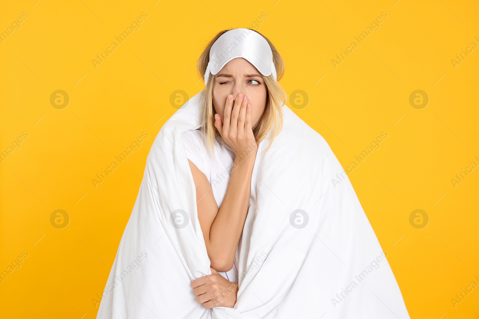 Photo of Young woman in sleeping mask wrapped with soft blanket yawning on yellow background