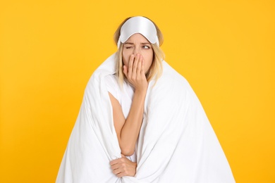 Young woman in sleeping mask wrapped with soft blanket yawning on yellow background
