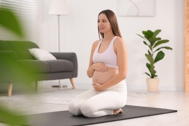 Pregnant woman sitting on yoga mat at home, space for text