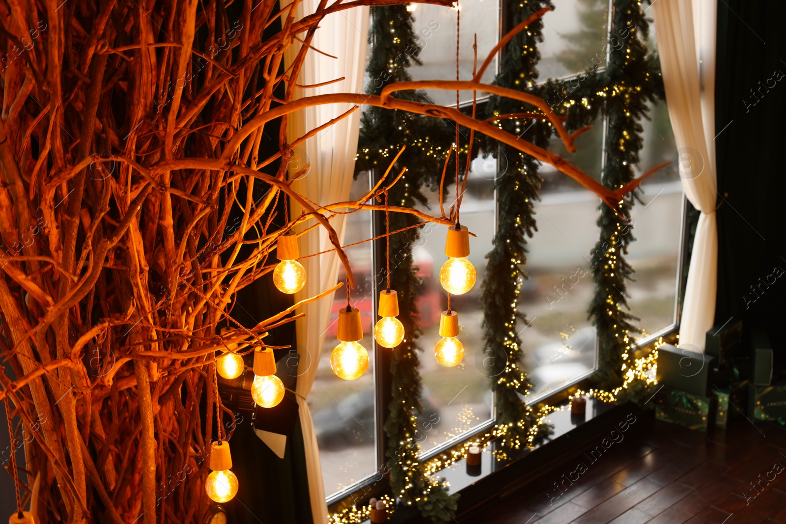 Photo of Window decorated with fir branches and Christmas lights and lamp bulbs hanging on dried tree in room