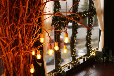 Photo of Window decorated with fir branches and Christmas lights and lamp bulbs hanging on dried tree in room
