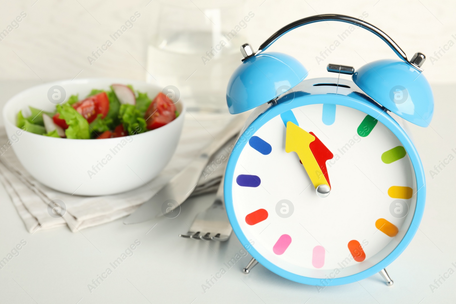 Photo of Alarm clock with salad on white table, closeup. Meal timing concept