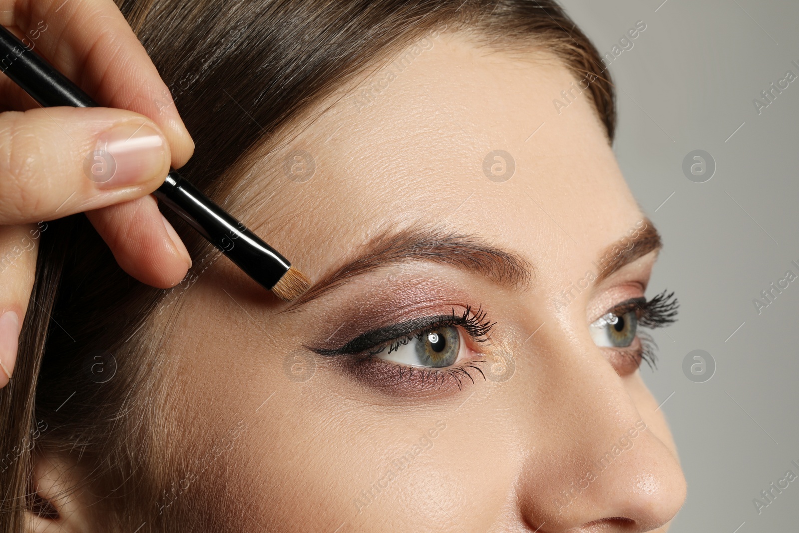 Photo of Artist correcting woman's eyebrow shape with powder on grey background, closeup